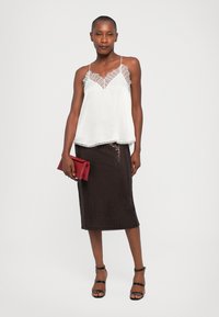 Camisole en soie blanche avec finition en dentelle, associé à une jupe crayon texturée noire, tenant une pochette rouge et portant des sandales noires à lanières.