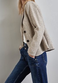 Blazer beige à motif pied-de-poule avec boutons noirs, associé à un jean en denim bleu foncé taille haute avec fermeture à boutons sur le devant.
