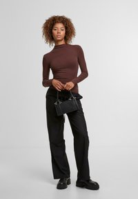 Braunes Langarmoberteil, kombiniert mit hochgeschnittenen schwarzen weit geschnittenen Hosen. Das Model hält eine kleine schwarze Handtasche mit silbernen Hardware-Details.