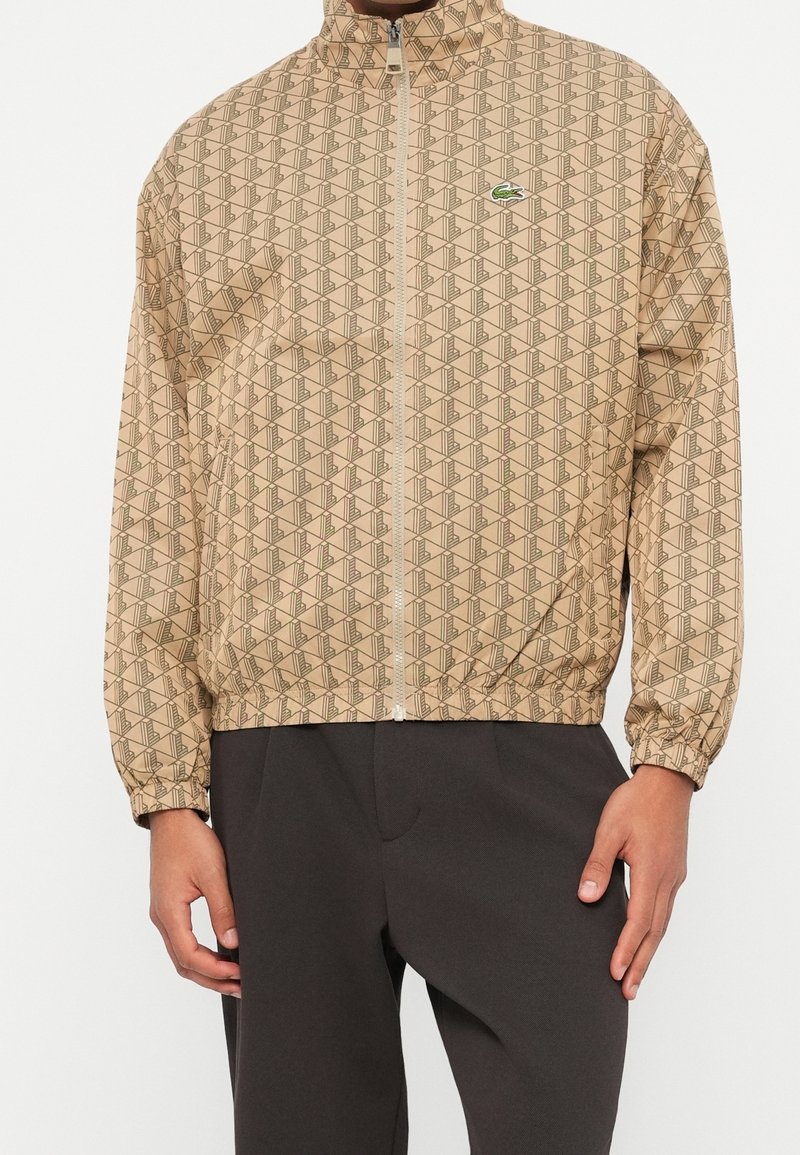Veste beige à motifs avec fermeture éclair, poignets élastiques et logo crocodile vert, portée avec un pantalon foncé par une personne debout.
