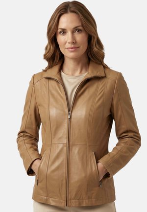 Frau trägt eine beige Lederjacke mit Reißverschluss und hat die Hände in den Taschen, steht vor einem schlichten weißen Hintergrund.