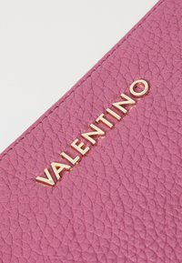 Rózsaszín textúrázott bőr, arany színű "VALENTINO" logóval debossálva. A közeli részlet kiemeli az anyag szemcsézettségét és a fémes kiegészítőket.