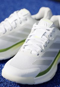 Ζευγάρι λευκά αθλητικά παπούτσια με λευκά κορδόνια, ανοιχτογκρι ρίγες και πράσινες λεπτομέρειες τοποθετημένα πάνω σε μπλε υφή.