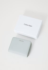 Ljust grå läderplånbok med dragkedja, präglad logotyp på framsidan, vilande på en vit låda med svart text, Calvin Klein.