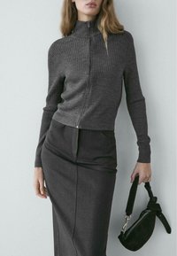 Grauer, gerippter Reißverschluss-Pullover mit hohem Kragen, kombiniert mit einem dunklen Bleistiftrock. Modell hält eine kleine schwarze Handtasche.