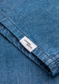 Tissu en denim de teinte bleue, présentant un bord plié et une étiquette blanche portant l'inscription "OMBRE WEAR". Coutures visibles le long de la couture.