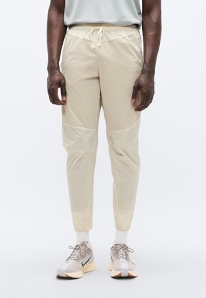 Lichtbeige sportbroek met een trekkoord in de taille, met een gladde textuur, taps toelopende pijpen en contrasterende zijpanelen. Gecombineerd met grijze sneakers.