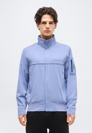 Jeune homme aux cheveux bouclés portant une veste à fermeture éclair bleu clair avec un col montant et une poche à rabat zippée sur la manche gauche.