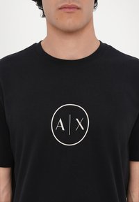 Sort bomulds t-shirt med et rundt grafisk logo med "A/X" i hvid. Korte ærmer og en rund halsudskæring med en glat tekstur.