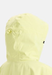 Casaco de chuva amarelo com capuz ajustado, textura suave e costuras subtis. Apresenta uma pequena etiqueta de logotipo preta na lateral.