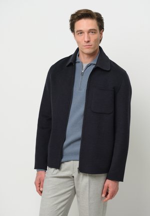 Dark navy zip-up jacket with a flat collar and front pocket, made from soft fabric. Worn over a light blue ribbed shirt and light gray pants.