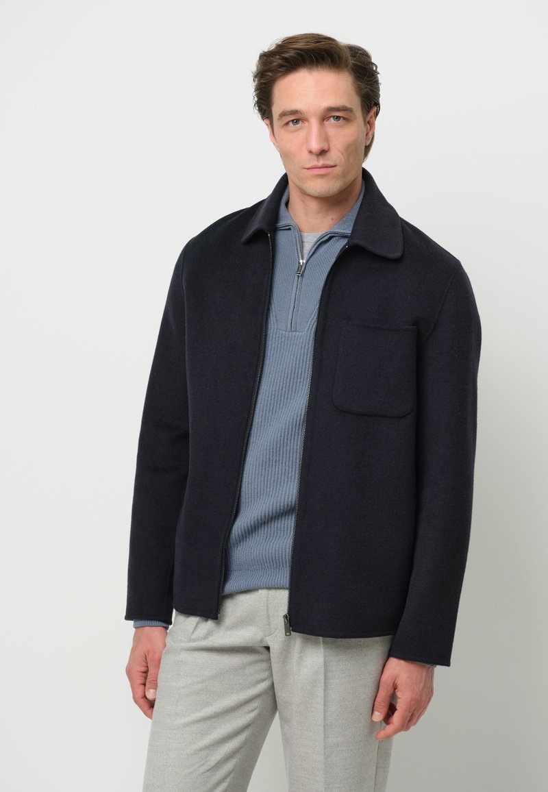 Giacca zip-up blu navy scuro con colletto piatto e tasca frontale, realizzata in tessuto morbido. Indossata sopra una maglietta a costine blu chiaro e pantaloni grigio chiaro.