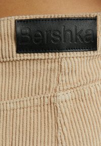Bežová kordurová látka s vertikálním žebrováním, s černou koženou etiketou s vyraženým nápisem "Bershka." Texturovaný povrch a čisté švy.