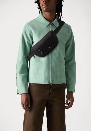 Svart bumbag båret over kroppen over en lys grønn suedjakke med glidelås og to frontlommer, kombinert med brune bukser.