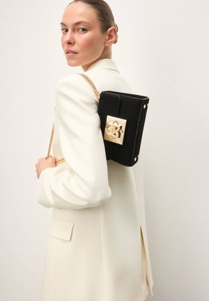 Czarna zamszowa torebka z złotym, geometrycznym zapięciem, z łańcuszkowym paskiem i prostokątnym kształtem. Noszona na boku, na tle jasnego płaszcza.