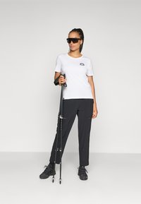 Weißes T-Shirt mit kurzen Ärmeln und einem Logo, kombiniert mit schwarzen Hosen und schwarzen Wanderschuhen. Die Person hält Trekkingstöcke und trägt Sonnenbrillen.