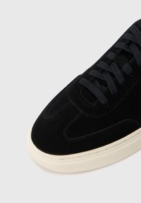 Svart semsket sneakers med svarte snører og hvit gummisåle, vist foran og delvis på oversiden på hvit bakgrunn.