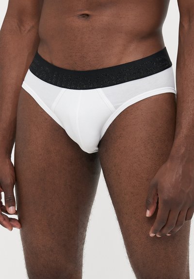 Hvide bomuldsunderbukser med en sort elastisk talje, der har et struktureret mønster. Stoffet virker blødt og let skinnende.