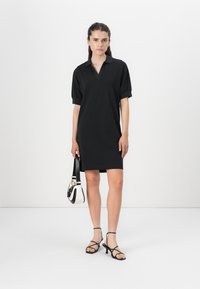 Black polo dress with short sleeves, made of soft fabric. Features a V-neck and a relaxed fit. Paired with black strappy sandals and a small bag.