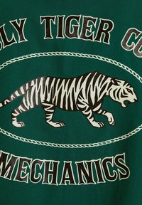 Tela verde con un gráfico de un tigre a rayas negro y blanco, rodeado por el texto "Holy Tiger Co. Mechanics" en negrita de color negro.