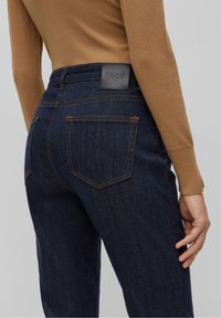 Mörkblå denimjeans med hög midja, som har orange sömmar, två bakfickor och en svart läder BOSS-tagg på midjebandet.