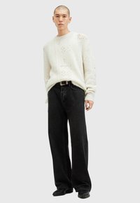 Cremefarbener Strickpullover mit offenen Mustern, kombiniert mit schwarzen weit geschnittenen Jeans. Das Model steht selbstbewusst mit natürlich positionierten Händen.