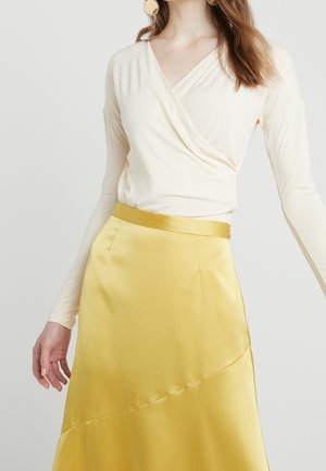 Femme portant un haut cache-cœur crème à manches longues associé à une jupe en satin jaune taille haute, montrée des épaules au milieu des cuisses sur un fond uni.