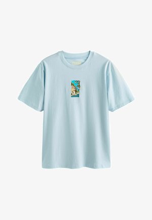 Camiseta de manga corta de color azul claro con un pequeño gráfico rectangular en el centro que muestra un paisaje costero con un acantilado, una casa y el océano.
