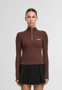 Braunes Langarmoberteil mit hohem Kragen und Reißverschluss, mit gerippter Textur und einem kleinen silbernen Akzent oberhalb der linken Brust. Schwarzer Plisseerock.