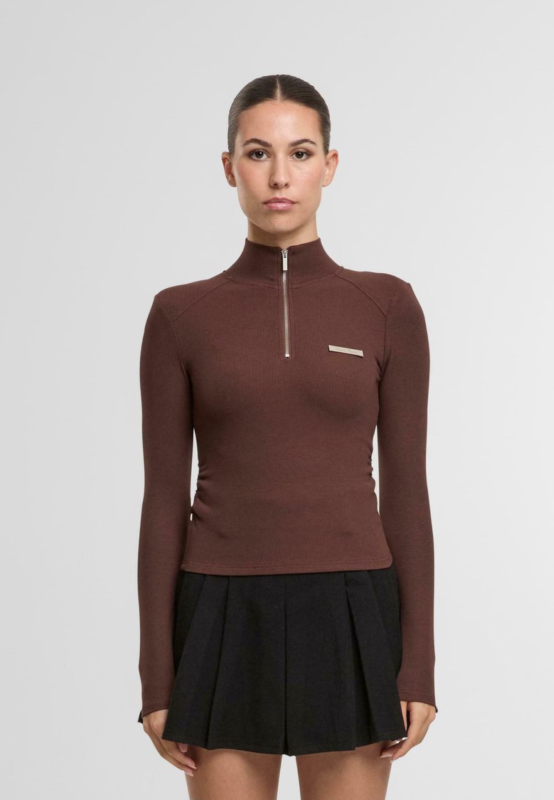 Braunes Langarmoberteil mit hohem Kragen und Reißverschluss, mit gerippter Textur und einem kleinen silbernen Akzent oberhalb der linken Brust. Schwarzer Plisseerock.