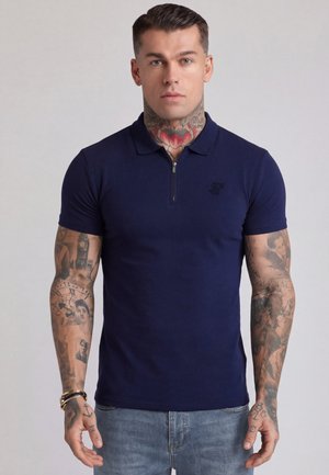 Muskuløs mand med hals- og arm-tatoveringer iført en marineblå kortærmet polo og lyseblå jeans, stående med fronten vendt fremad.