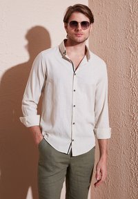 Camisa de lino beige claro con un collar estructurado, botones negros y mangas enrolladas, combinada con pantalones verde oliva.