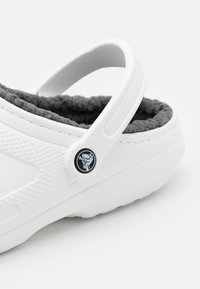 Sabot blanc avec une doublure en peluche grise, un extérieur texturé et un accent logo circulaire noir. Dispose d'une sangle au talon ajustable pour un maintien sécurisé.