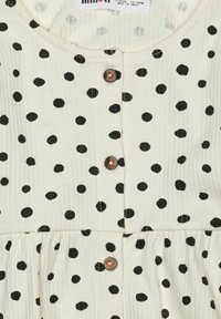 Bílá látka šatů s černými puntíky, s kulatým výstřihem a třemi hnědými knoflíky vpředu. Viditelný plisovaný design.