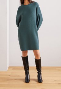 Vestito a maglia teal a coste con vestibilità ampia e maniche lunghe, abbinato a stivali neri in pelle al ginocchio, su un pavimento in legno chiaro.