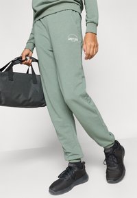 Zielone spodnie dresowe z elastycznym pasem, bocznymi kieszeniami oraz białym nadrukiem "LIMITLESS". W zestawie z czarnymi trampkami i trzymającą czarną torbę sportową.