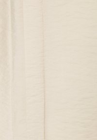 Tessuto beige testurizzato con sottili ondulazioni, con cuciture visibili che creano un effetto a strati. Il materiale appare leggero e morbido.