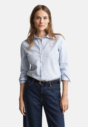 Light blue button-up shirt with long sleeves, featuring a small logo near the chest, paired with dark blue denim jeans and a black belt.