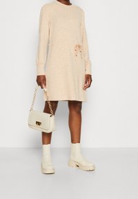 Robe en maille pêche à manches longues avec ceinture nouée à la taille, associée à des bottines beiges épaisses et un sac à main crème avec des éléments dorés.