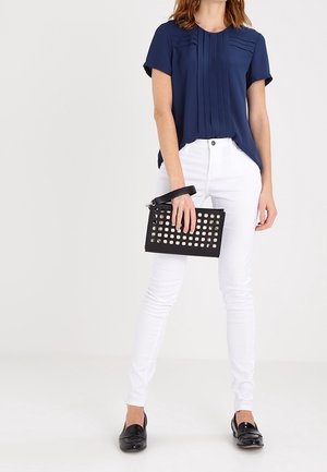 Marineblauwe blouse met korte mouwen en verticale plooien, witte slim-fit jeans en een zwarte geperforeerde clutch; zwarte loafers maken de outfit compleet.