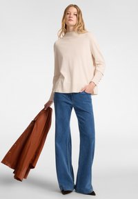 Vaaleanbeige neulepusero, jossa on korkea kaulus ja ribbireunat, yhdistetty tummansinisiin leveälahkeisiin farkkuihin. Vasemmassa kädessä pidetään ruskeaa nahkatakkia.