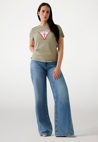 T-shirt verde oliva con logo triangolare rosso e bianco; abbinato a jeans larghi di colore azzurro chiaro con una texture denim classica.