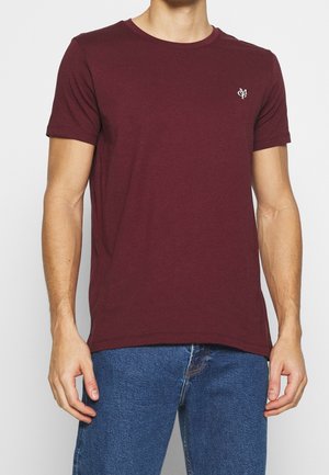Mann trägt ein schlichtes dunkelrotes T-Shirt mit kurzen Ärmeln und einem kleinen weißen Logo auf der linken Brust sowie blaue Jeans, steht vor einem weißen Hintergrund.