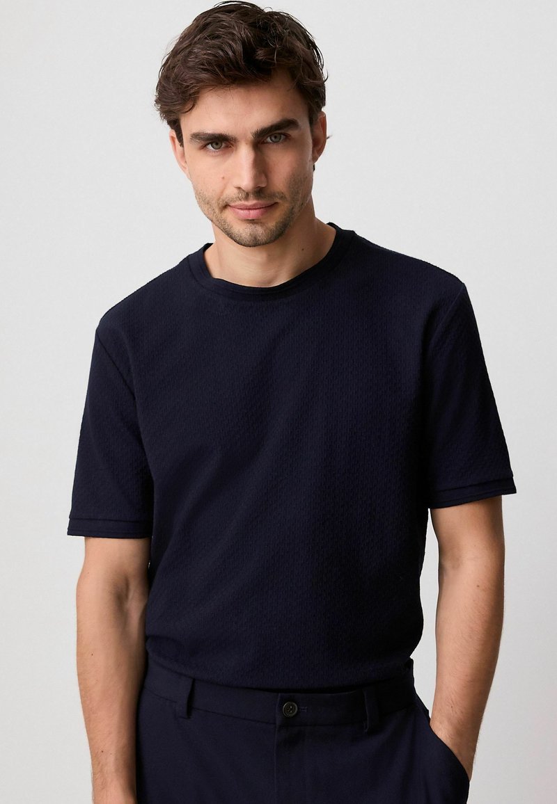 T-shirt bleu marine texturé à manches courtes avec un col rond, présentant un motif subtil et une coupe décontractée, associé à un pantalon assorti.