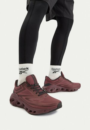 Persona che indossa scarpe da corsa Reebok rosso scuro, calzini Reebok bianchi e leggings neri, in piedi contro uno sfondo chiaro e semplice.