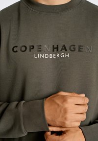 Sudadera verde oscuro con una textura de canalé, que presenta el texto en negrita y en relieve "COPENHAGEN" en negro y "LINDBERGH" en blanco.