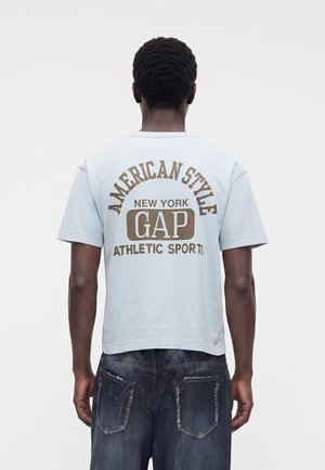 Osoba chrbtom, oblečená v svetlomodrom tričku s "American Style New York GAP Athletic Sports" v hnedom potlači a vyblednutých tmavých džínsoch.