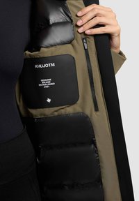 Isolierte Jacke mit khakifarbenem Außenmaterial, schwarzem Pufferfutter und einer Reißverschlusstasche, die mit einem Markenpatch und Logo versehen ist. Strukturierter Stoff bildet einen Kontrast zu den glatten Abschnitten.