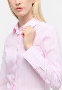 Camicia a maniche lunghe di colore rosa chiaro con righe verticali bianche. Presenta un colletto, bottoni ai polsini e una texture di tessuto liscia.