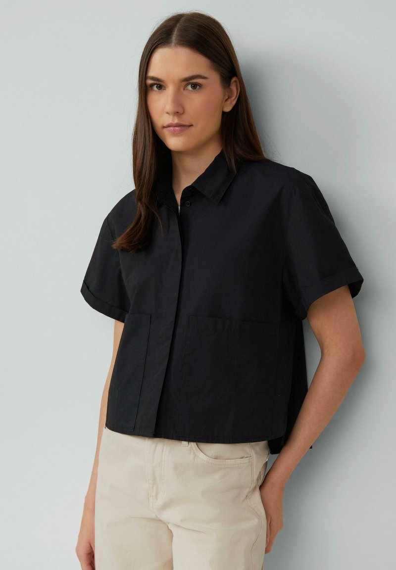 Chemise noire à manches courtes avec col, dotée de deux poches avant et d'une coupe décontractée. Le tissu semble lisse et respirant.